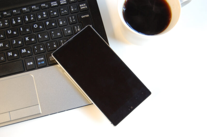 白いテーブルの上に、ノートパソコン、スマートフォン、コーヒーが乗っている画像